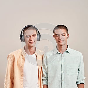 Two young caucasian twin brothers in casual wear listening music in headphones and wireless earphones, while standing