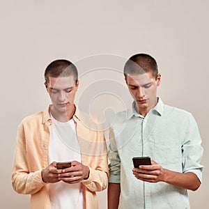 Two young caucasian twin brothers in casual wear using smartphones while standing isolated over beige background