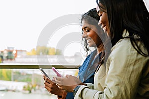 Two young cheerful multinational women using cellphones while walking outdoors