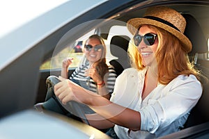 Two young cheerful smiling women in a car on vacation trip to the sea beach. Girl in glasses driving a vehicle from