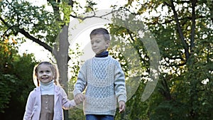 Two young children, an older boy and a younger girl, are walking in the park, holding hands. The children are in high