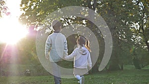 Two young children, an older boy and a younger girl, are walking in the park, holding hands. Sunset. Rear view.