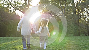 Two young children, an older boy and a younger girl, are walking in the park, holding hands. Sunset. Rear view.