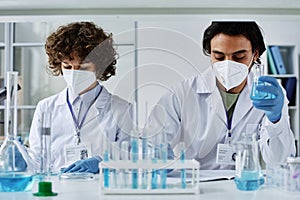 Two young clinicians in labcoats, respirators and gloves working with liquids