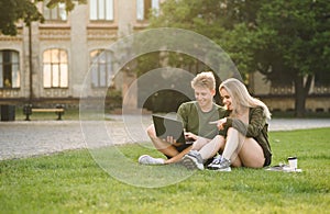 Two young college students watching funny video on the laptop, preparing for the exam with computer sitting on the lawn. Couple of