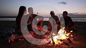 Two young couples are sitting on the beach near the bonfire. Roasting meat on wooden sticks, eating. Happy time together