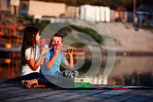 Two young cute little friends, boy and girl talking, drinking tea, eating sandwiches and fishing on a lake in a sunny summer day