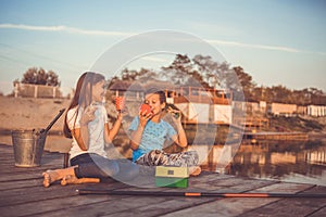 Two young cute little friends, boy and girl talking, drinking tea, eating sandwiches and fishing on a lake in a sunny summer day