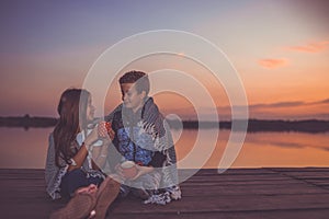 Two young cute little friends, boy and girl talking and drinking tea while sitting covered with blanket. sunset