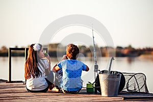 Two young cute little friends, boy and girl talking, eating sandwiches and fishing on a lake in a sunny summer day