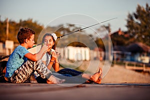 Two young cute little friends, boy and girl talking, eating sandwiches and fishing on a lake in a sunny summer day