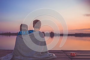 Two young cute little friends, boy and girl talking while sitting hugging and covered with blanket by the lake during sunset