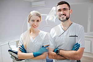 Two young dentist professionals in dentistry room. They pose on camer and smile. People hold hands crossed. photo