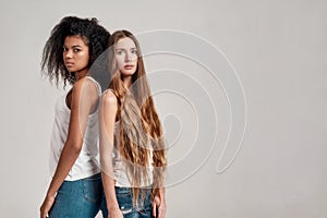 Two young diverse women wearing white shirts looking at camera while standing together isolated over grey background
