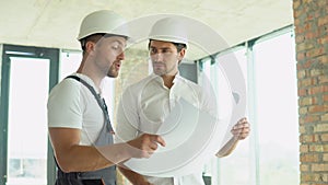 Two young engineer in a safety helmets standing at building site holding a blueprint of the structure. Construction