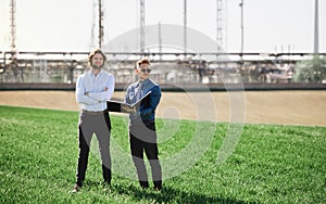 Two young engineers standing outdoors by oil refinery, discussing issues.