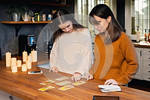 Two young femaies playing with tarot cards in a kitchen. Candles and cellphones visible