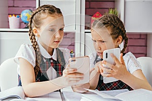 Two young female classmates are sitting at a desk and using smartphones. Modern technology concept