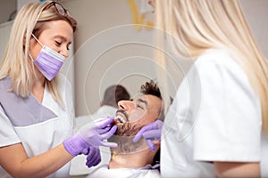 Two young female dentists working in dental clinic. Whitening male patient`s teeth and using tone chart to mach color photo