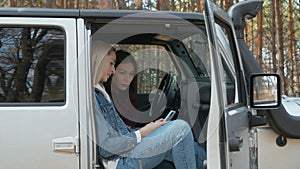 Two young female friends sits in a white car jeep and uses phone in autumn forest
