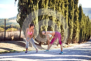 Two young female friends stretching, worming up before jogging, looking each other, smiling