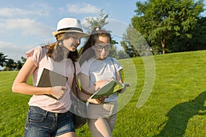 Two young female students with backpacks, books go against the background of green lawn, park.