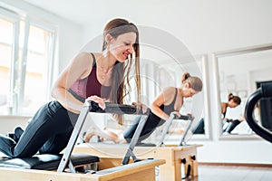 Two young females doing ABS muscles strength exercises using pilates reformer machine in sport athletic gym hall. Active people