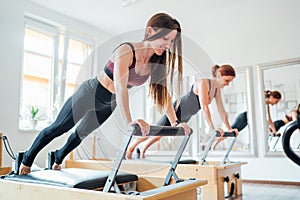 Two young females doing extended plank static strengthening core muscles exercise using pilates reformer machine in sport athletic