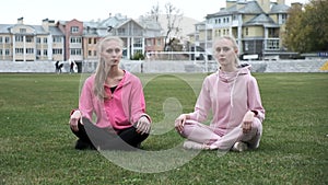 two young fit twin sisters in pink sportswear sit at grass field in lotus pose