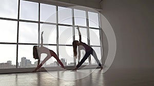 Two young fit women practicing yoga poses synchronously standing against window background