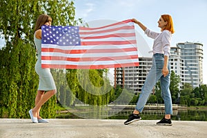 Two young friends women holding USA national flag in their hands standing together outdoors. Patriotic girls celebrating United