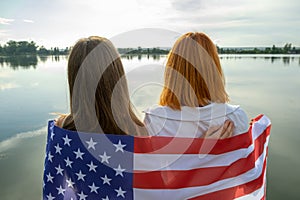 Two young friends women with USA national flag on their shoulders standing together outdoors on lake shore. Patriotic girls