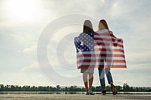 Two young friends women with USA national flag on their shoulders standing together outdoors on lake shore. Patriotic girls