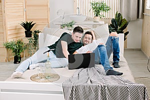 Two young gay men lying on the bed and using a laptop. Handsome gay men spending time together. Homosexual couple, gay