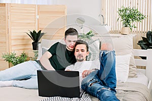 Two young gay men lying on the bed and using a laptop. Handsome gay men spending time together. Homosexual couple, gay