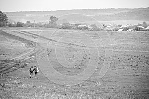 Two young girls with backpacks and photo camera make selfie on phone near rural road. Horse farm pasture with mare and foal.