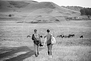 Two young girls with backpacks and photo camera on rural road. Horse farm pasture with mare and foal. Small village with old house