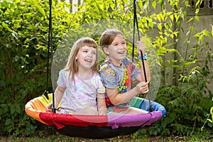 Two young girls, children sitting side by side on a round swing, joyful happy cheerful kids laughing together, sisters siblings,