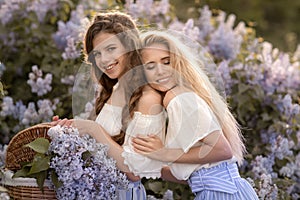 Two young girls dressed in beautiful clothes stand against the background of lilac bushes