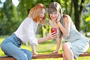 Two young girls friends sitting on a bench in summer park looking in red smartphone