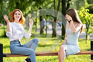 Two young girls friends sitting on a bench in summer park talking happily having fun