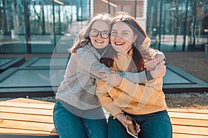 Two young girls friends smiling and hugging while sitting in a modern place looking at the camera