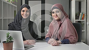 Two young girls in hijab sit in the office with a look at the camera. Smiling, positive 60 fps