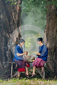 Two young girls prepare food using motar and other flavoring look like to make papaya salad and they sit under big tree with day