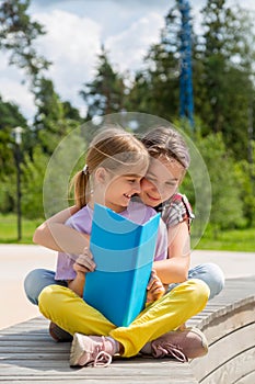 Two young girls sitting on the bench outdoors reading a book.