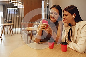 Two young girls are sitting in cafe with modern interior. Smiling longhaired brunettes women looking at smartphone at restaurant