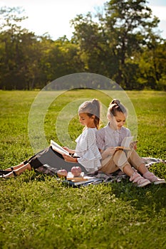 Two young girls sitting on green grass outdoors reading separate books. Friends enjoying time together in summer in park