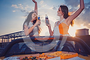 Two young girls smiling, cheering with soda in glass bottles, posing in yellow car cabrio with french fries and pizza on