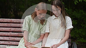 Two young girls in summer dresses sit on wooden bench and read book in park. School girls learning lessons and preparing