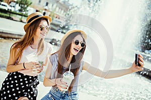 Two young girls in sunglasses and hats near city fountain. Best friends drink cocktails doing self photo and laughing in city stre